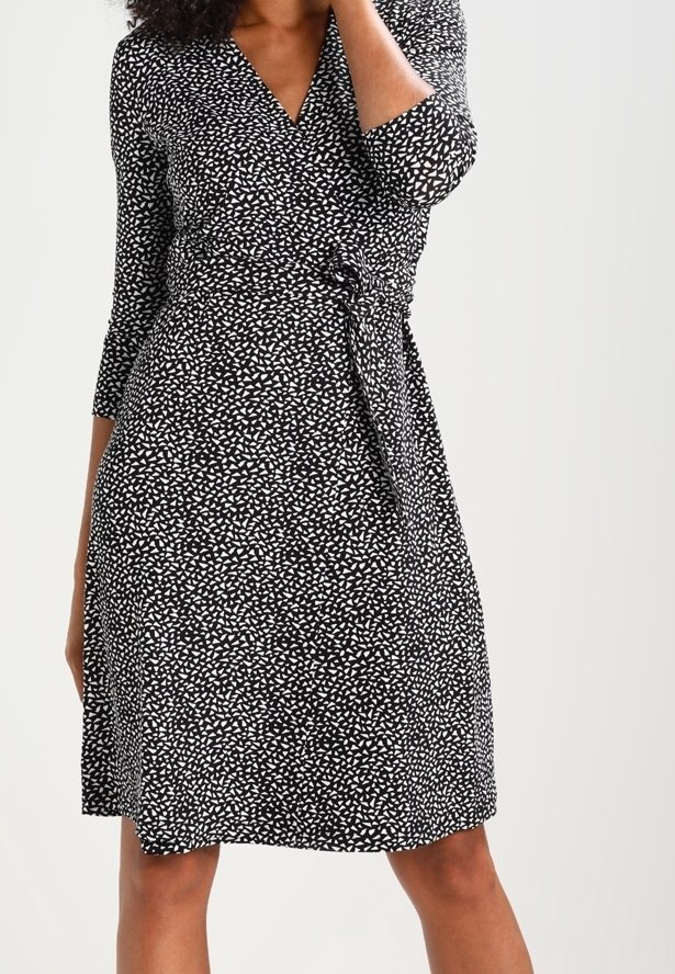 Femme portant une robe portefeuille noire avec un motif abstrait de triangles blancs et des manches trois-quarts, se tenant avec une main près du visage.