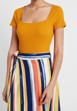 Femme portant un haut à manches courtes de couleur jaune moutarde avec un col carré, et une jupe à rayures verticales multicolores avec des bandes bleu marine, orange, jaune et blanches.