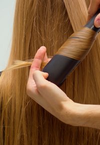 Hånd, der bruger en roterende varmluftsbørste til at krølle en sektion af langt, glat, blondt hår mod en ensfarvet baggrund.