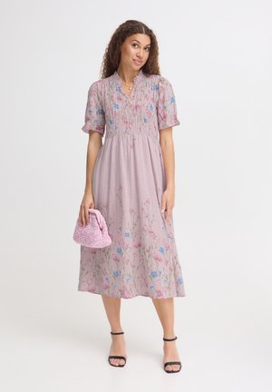 Kvinde iført en lyserød blomsterprintet midi-kjole med puffede ærmer, der holder en tekstureret lyserød håndtaske og har sorte sandaler med ankelrem på en hvid baggrund.