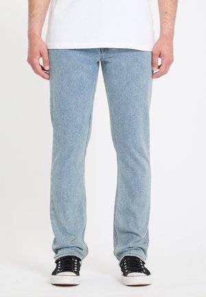 Homme portant un jean droit bleu clair, des baskets basses noires et une chemise blanche devant un fond uni.