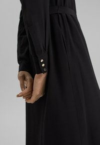 Długa czarna sukienka z dopasowaną talią i guzikami przy mankietach; charakteryzuje się gładką teksturą i elegancką sylwetką, podkreślającą prostą elegancję.