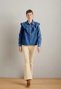 Τζιν τοπ με μεγάλο ρυτιδωτό γιακά, κλείνει μπροστά με κουμπιά. Μπεζ παντελόνια με καμπάνα, καφέ μπότες. Λείος υφασμάτινος χαρακτήρας, χαλαρή εφαρμογή. Ουδέτερο φόντο.