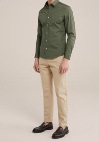 Mężczyzna ubrany w oliwkową koszulę na guziki, jasnobeżowe spodnie i ciemnobrązowe loafersy, stojący na jednolitym beżowym tle.