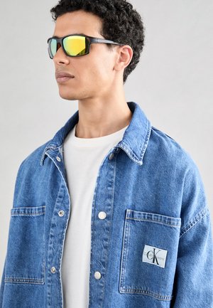 Modrá džínsová bunda s knoflíkovým zapínáním, dvěma předními kapsami a logem. Pod ní je bílá tričko, stylizované s černými slunečními brýlemi.