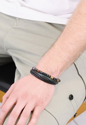 Leather and bead bracelets in black, green, and earth tones, featuring braided and smooth textures, worn on a wrist on a beige fabric surface.