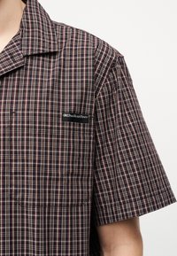 Πουκάμισο κοντομάνικο μεσίστυλο σε σκούρο καρό με γιακά και τσέπη στο στήθος που διαθέτει μαύρο έμπλαστρο με την ετικέτα "Quiksilver" σε ανοιχτό φόντο.