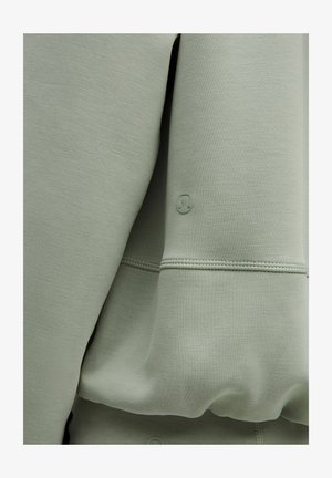 Grøn sweatshirt med en glat, blød tekstur, synlig syning og en lille logo detalje på ærmet.