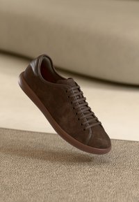 Zapatillas deportivas de ante marrón con una suela de goma lisa, que presentan cordones redondos y un acabado texturizado, suspendidas sobre una superficie beige.