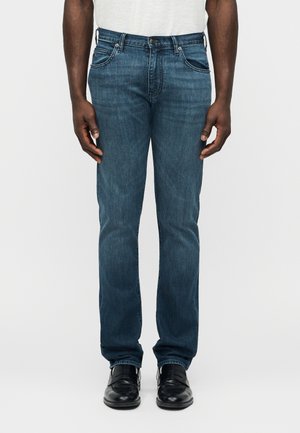 Täiskasvanu kannab keskmise sinise värviga sirge säärega teksapükse, musti nahast jalanõusid ja valget särk heledal ühevärvilisel taustal.