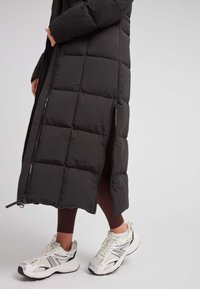 Abrigo largo negro tipo plumón con un diseño acolchado, que presenta cremalleras laterales. Combinado con zapatillas deportivas blancas y plateadas y leggins marrones.