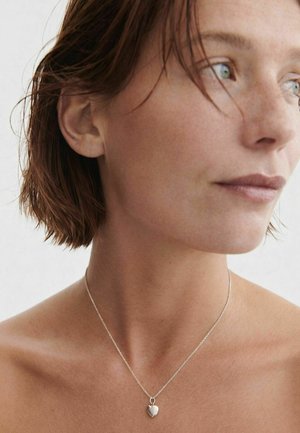 Kobieta o krótkich brązowych włosach, nosząca srebrny naszyjnik z wisiorkiem w kształcie serca, spoglądająca w bok na jednolitym tle.