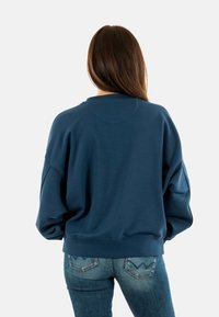 Sudadera de gran tamaño en azul oscuro con hombros caídos, cuello de canalé y detalles en los puños; combinada con jeans de mezclilla azul claro.
