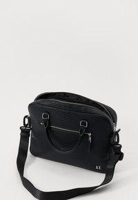 Sacoche d'ordinateur portable noire en matériau texturé, avec des poignées doubles, un compartiment zippé, et une sangle détachable avec un détail de fermeture éclair en ton argenté.