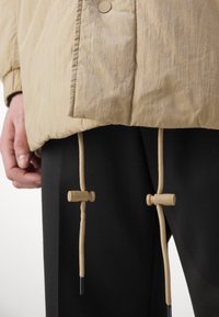 Beige pufferjack met een gestructureerde stof, drukknoopjes en verstelbare trekkoorden in zandtinten, gecombineerd met zwarte broeken.