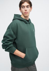 Sweatshirt verde em tecido suave com bolsos canguru, punhos canelados e um logo discreto no peito. Design simples, corte descontraído, comprimento médio.