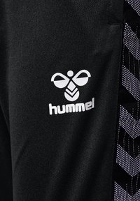 Czarne spodnie sportowe z wyraźnym białym logo na udzie. Boczne panele zdobi geometryczny wzór w odcieniach czerni i fioletu.