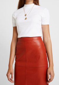 Bílý žebrovaný top s krátkým rukávem a vysokým límcem, který je spojen s těsnou červenou koženou sukní se švy. Dvě vrstvené zlaté náhrdelníky.