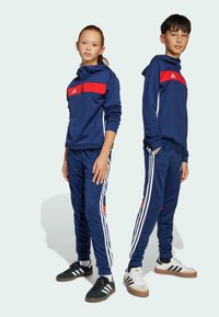 Adidas Trainingsanzüge in Marineblau mit roten Akzenten. Die Kapuzenoberteile und die passenden Hosen haben weiße Streifen an den Seiten. Der Stoff wirkt glatt und eng anliegend.