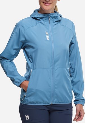 Femme portant une veste à capuche zippée Millet bleu clair et un pantalon bleu marine assorti, les mains dans les poches de la veste, sur fond blanc