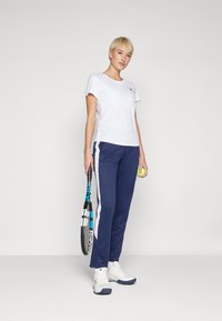 Hvit kortermet treningstrøye, marineblå joggebukser med sidestriper, hvite joggesko, holder en tenniskølle og ball. Enkel, funksjonell design.