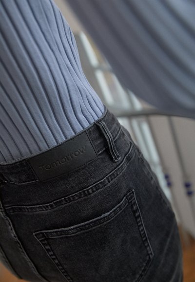 Jeans de ganga pretos com uma textura suave, apresentando um rótulo retangular preto com a inscrição "Tomorrow" na cintura e uma blusa de tom azul claro com nervuras.