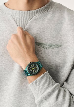 Ceas digital turcoaz cu cadran negru și accente galbene, purtat pe încheietura mâinii. Hanorac gri cu guler texturat și logo subtil.