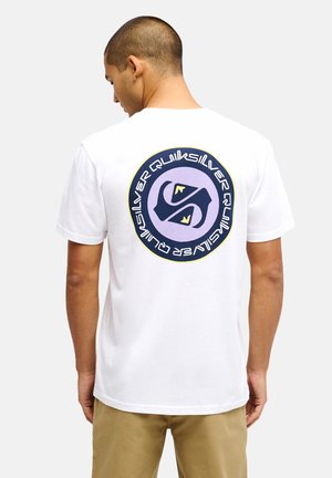 Mężczyzna w białej koszulce z dużym okrągłym logo Quiksilver w kolorach fioletowym, granatowym i żółtym na plecach, w połączeniu z beżowymi spodniami.