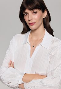 Biała, pomarszczona bluzka z kołnierzykiem i zapięciem na guziki przy mankietach. Złoty naszyjnik z ciemnym wisiorem. Modelka z włosami w średnim odcieniu brązu.