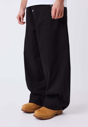 Homme portant un pantalon noir ample et des bottes en daim beige, debout sur une surface blanche, jambes inférieures et mains visibles.