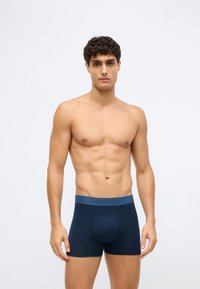 Young man with dark curly hair wearing navy blue boxer briefs with a matching waistband, standing against a plain light background.