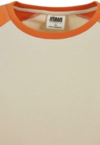Ekologiška medvilninė marškinėlių su grietinėlės spalvos kūnu ir oranžiniais raglan rankovėmis. Yra įrengtas banguotas apykaklė ir etiketė, rodanti miesto klasikų prekės ženklą.