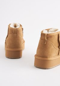 Brune ruskindsankelstøvler med blødt hvidt for, flad sål og lynlåsdetaile på siden. Har en polstret hæl for komfort.