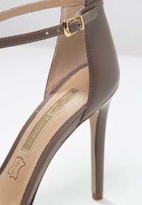 Sandale à talons hauts marron avec une tige en cuir brillant, une sangle pour la cheville ornée d'une boucle dorée, et un intérieur beige contrastant.