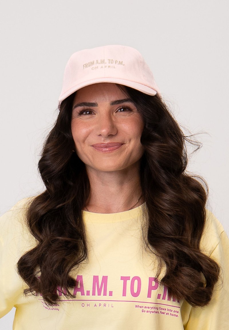 Femme aux longs cheveux bruns ondulés, portant une casquette rose clair et un t-shirt jaune avec un texte rose, souriant devant un fond uni.
