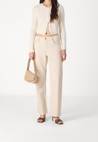 Cardigan corto crema, pantaloni beige a gamba larga, borsa beige e sandali con tacco marroni con accenti borchiati. Texture morbide e colori neutri.