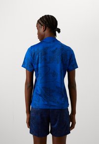 Blue athletic shirt with black abstract pattern, short sleeves, and a collar, paired with dark patterned shorts. Smooth, breathable fabric.