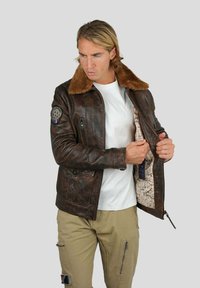 Veste en cuir marron avec un col en fausse fourrure, poches zippées et doublure intérieure à motifs, portée sur une chemise blanche et un pantalon beige.