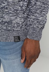 Stickad tröja med en melerad grå och marinblå design, ribbade ärmslut och en etikett med vit text vid nederkanten. Kombinerat med ljusblå jeans.