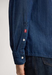 Camicia in denim di colore blu scuro, con un'etichetta rossa con punto esclamativo e dettagli in pulsante argentato sui polsini.