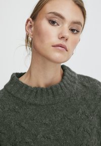 Dunkelgrüner Strickpullover mit hohem, geripptem Kragen und strukturiertem Muster. Die Haut hat einen natürlichen Farbton; die Ohrringe sind klein und in Goldton.