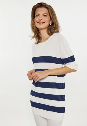 Kvinde smilende iført en hvid sweater med brede mørkeblå vandrette striber, stående med hænderne foldet foran en ensfarvet hvid baggrund.