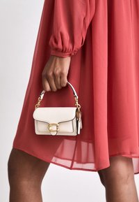 Borsa a tracolla bianca in pelle con una chiusura dorata, manico corto e rifiniture nere, tenuta in mano davanti a un abito rosso fluente.