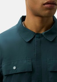 Turkusowa, lekka koszula z kołnierzykiem i dwoma kieszeniami na piersi. Posiada zapięcie na guziki oraz fakturowaną tkaninę.