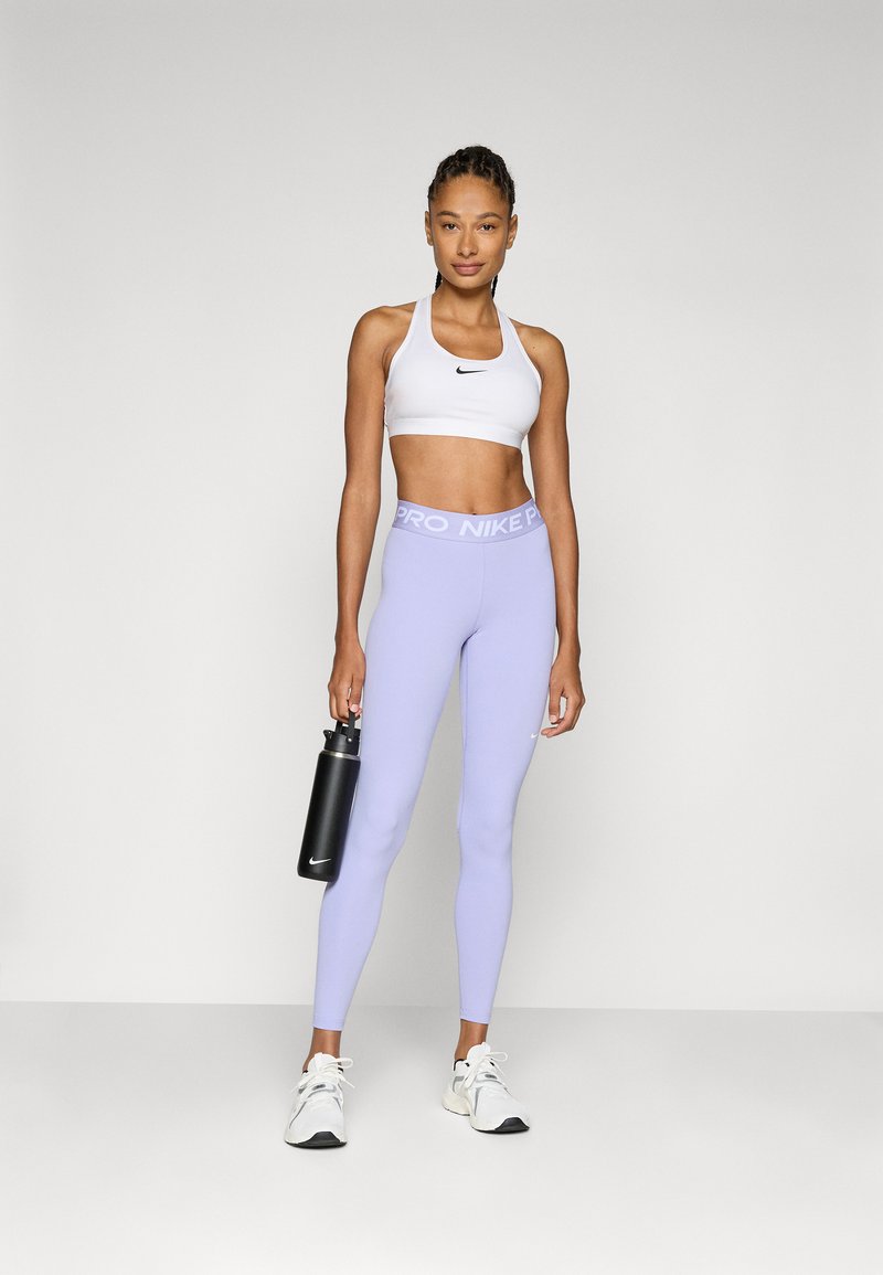 Sujetador deportivo blanco con logotipo negro de swoosh, leggings morados claros con cinturilla "PRO NIKE", botella de agua negra y zapatillas deportivas blancas.