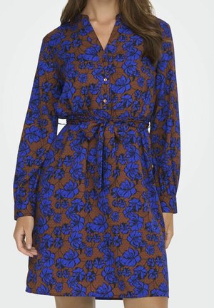 Woman wearing knee-length, long-sleeve brown dress with blue floral pattern, buttoned front, and tied waist belt against plain background.