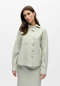 Chemise gris clair boutonnée avec col, manches longues, poche poitrine et motif texturé subtil. Jupe assortie visible en bas.