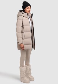 Jachetă puffer bej cu glugă, cu fermoar și buzunare laterale. Asortată cu colanți ușor bej cu model și cizme de iarnă cu toc.