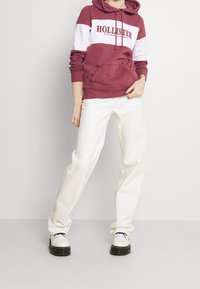 Persona vistiendo una sudadera burgundy y blanca de Hollister, pantalones rectos en color blanco roto y zapatos lace-up blancos, de pie contra un fondo liso.