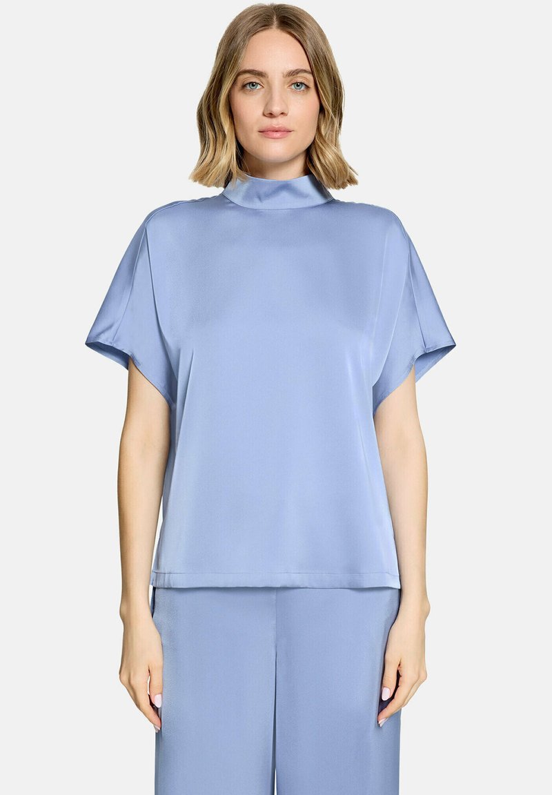 Femme portant un haut ample bleu clair à manches courtes avec un col montant et un pantalon large assorti, debout devant un fond uni.
